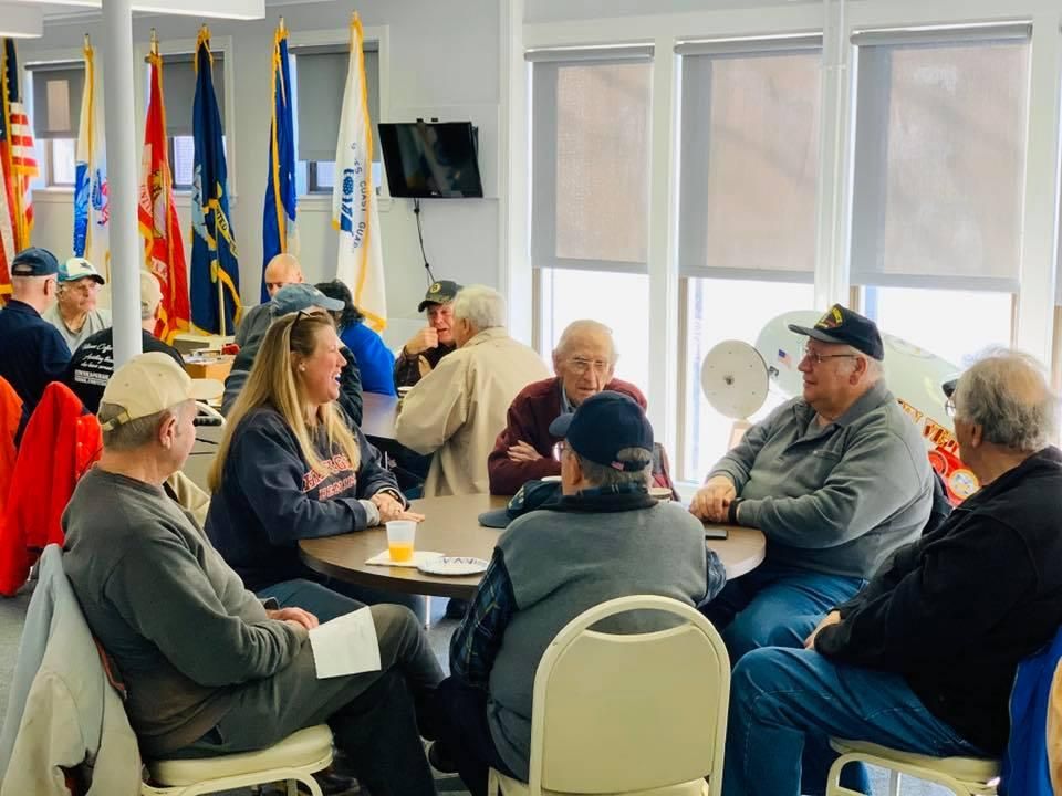 Veterans and volunteers gathered around tables at a community fundraising event, service flags along the wall